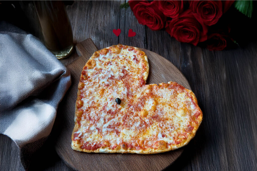 Heart-shaped cheese pizza on a wooden board, garnished with a single olive, surrounded by red roses and romantic decor.